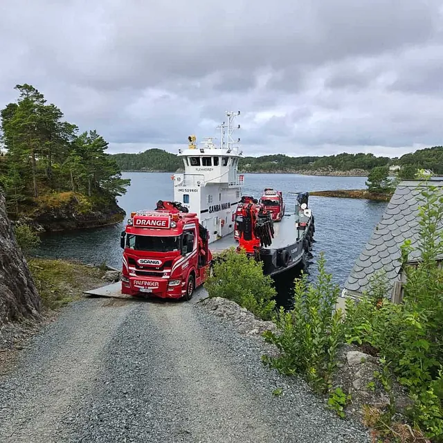 Rød lastebil kjører av en ferge på en grusvei ved sjøen.