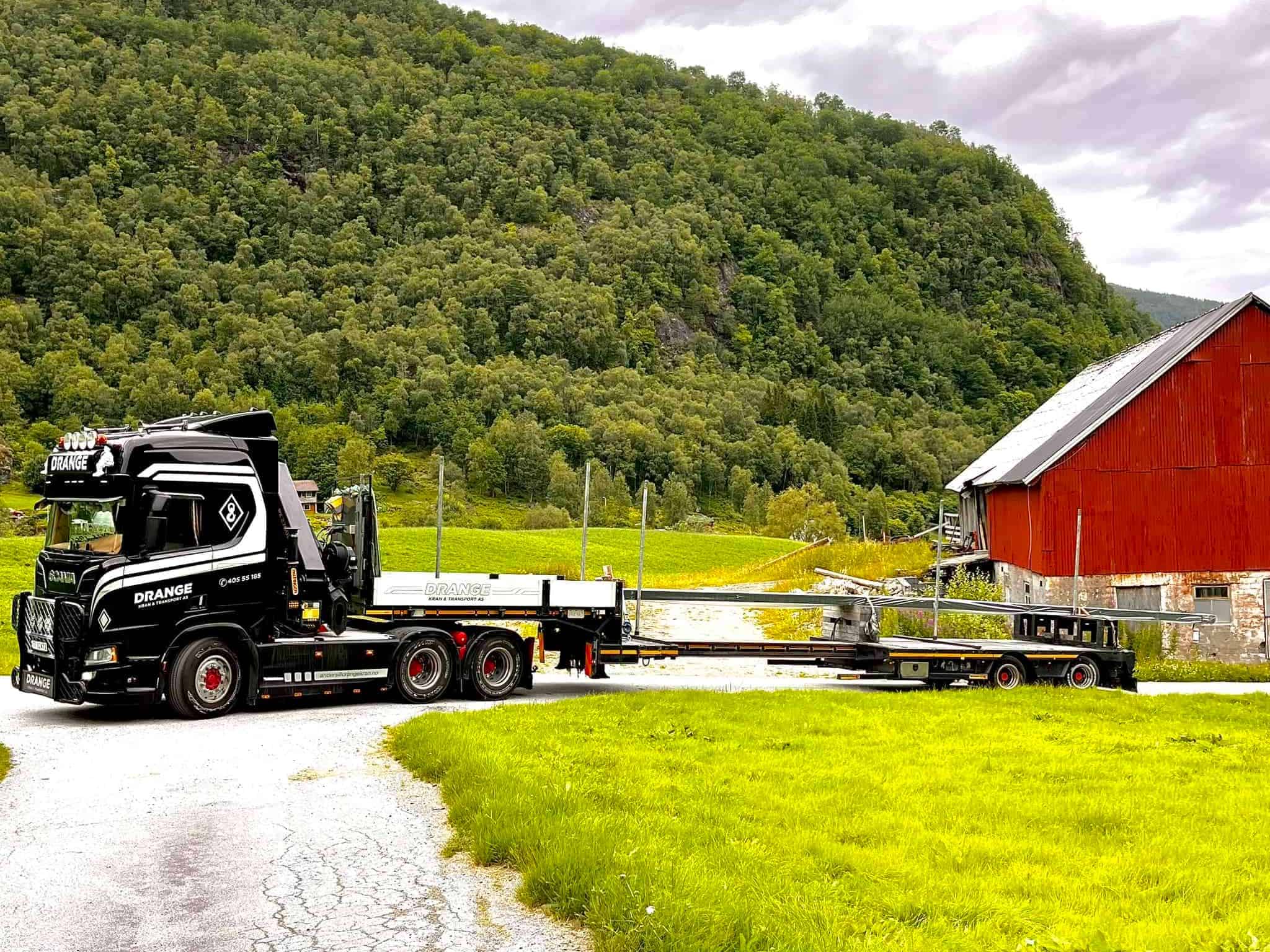 Drange lastebil på landevei med fjell i bakgrunnen.