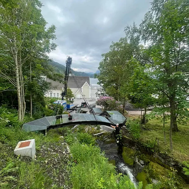 Metallbro monteres over en liten elv i grønn natur.