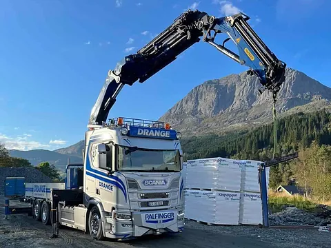 Hvit og blå Drange lastebil med kranarm på en grusplass med et fjell i bakgrunnen