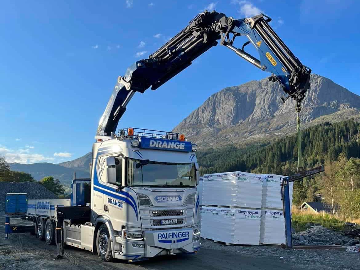 Hvit og blå Drange lastebil med kranarm på en grusplass med et fjell i bakgrunnen