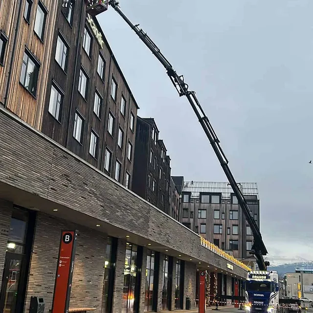 Kranbil løfter en arbeider i kurv opp til hotellfasade.