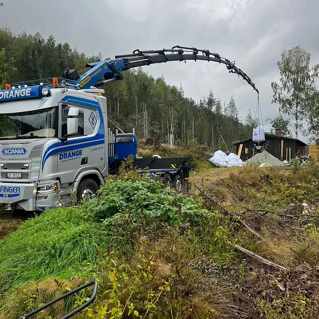 Kranbil løfter en storsekk ved en liten hytte i skogen.